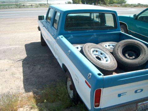 1978 Ford Bronco - A two-tone Ford pickup truck is parked on a dirt lot next to a highway, carrying spare tires in its bed.