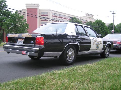1991 Ford LTD Crown Victoria - Rear view of a black-and-white Ford Highway Patrol sedan with star shield parked at a curb in front of a tan building.
