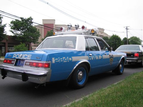 1991 Ford LTD Crown Victoria - Rear quarter view of a blue-and-white NYPD sedan marked POLICE 2840 HWY 3 with multiple roof lights parked at a curb.