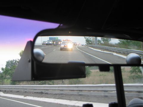 1991 Ford LTD Crown Victoria - Reflected in a rearview mirror at dusk, a police car with headlights on follows the camera vehicle along a guardrail-lined highway.