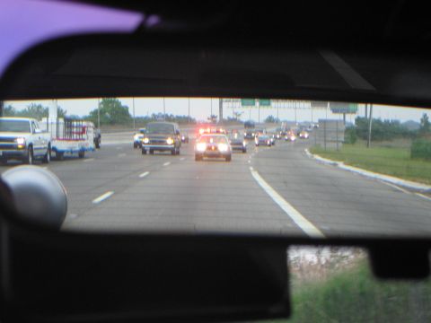 1991 Ford LTD Crown Victoria - View through a rear window of highway traffic, a police car with red roof lights and a pickup hauling a trailer among the cars.