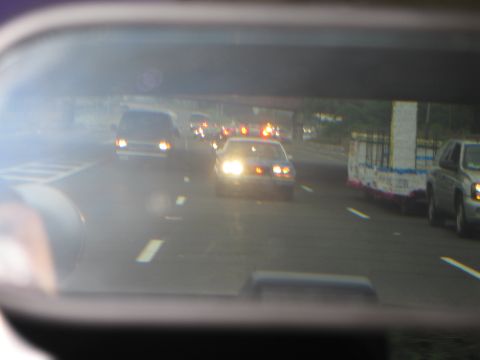 1991 Ford LTD Crown Victoria - Reflected in a side mirror, a police car with headlights on travels under a highway overpass amid other vehicles.