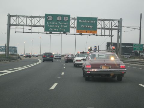 1991 Ford LTD Crown Victoria - maroon sedan with a roof light leads a small convoy on a highway beneath green I-495 Lincoln Tunnel and Meadowland Parkway signs.