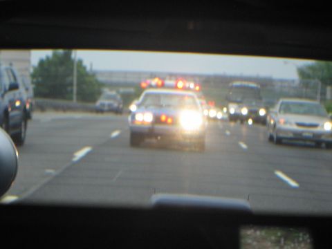 1991 Ford LTD Crown Victoria - Through a windshield, an oncoming police car runs flashing emergency lights amid traffic on a wide highway at dusk.