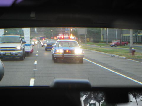 1991 Ford LTD Crown Victoria - police car with red roof lights leads a line of vehicles down a city roadway, viewed through a windshield.
