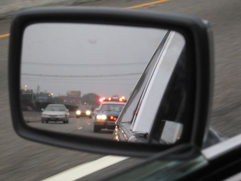 1991 Ford LTD Crown Victoria - car's side mirror reflects a police vehicle with red emergency lights following on a misty highway.