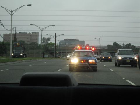1991 Ford LTD Crown Victoria - police car runs flashing red lights down a broad roadway lined with power lines and distant buildings under gray skies.