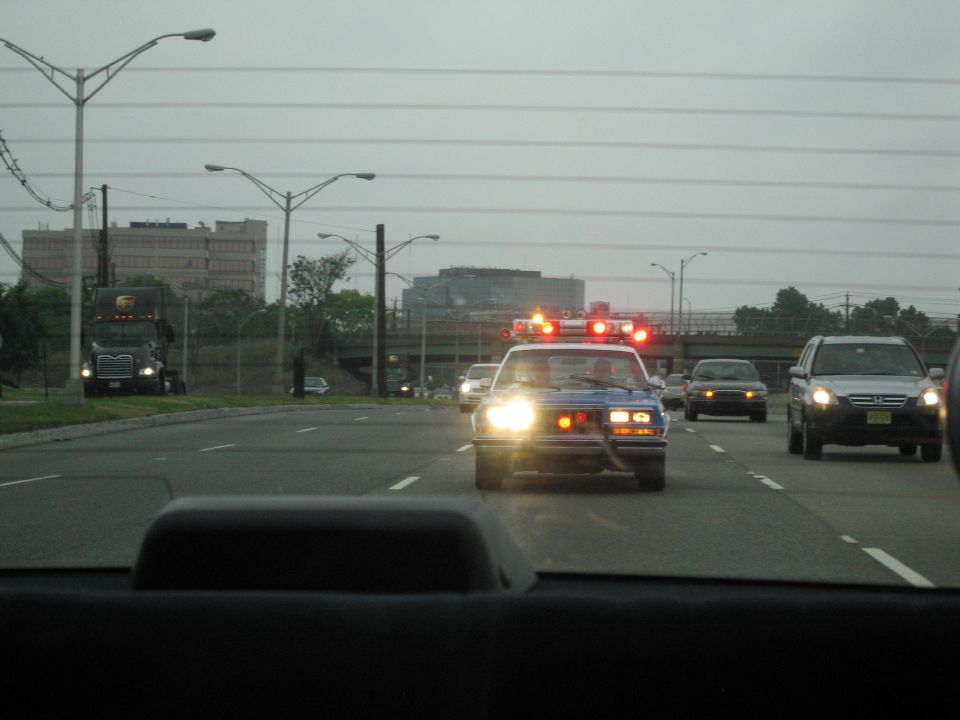 Police car with flashing red lights on a wide road with power lines and city buildings