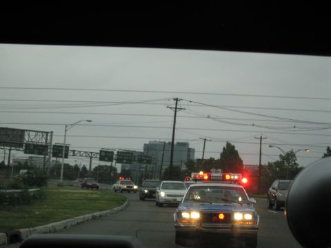 1991 Ford LTD Crown Victoria - police car flashes red and amber emergency lights at the roadside near a highway interchange under overcast skies.