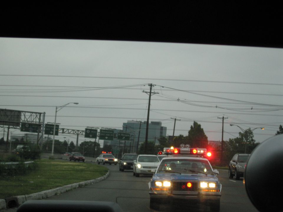 Police car with red and orange emergency lights at a roadside near a highway interchange