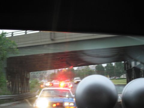 1991 Ford LTD Crown Victoria - Through a dark windshield, a police car with bright headlights and red flashers passes beneath a highway overpass.