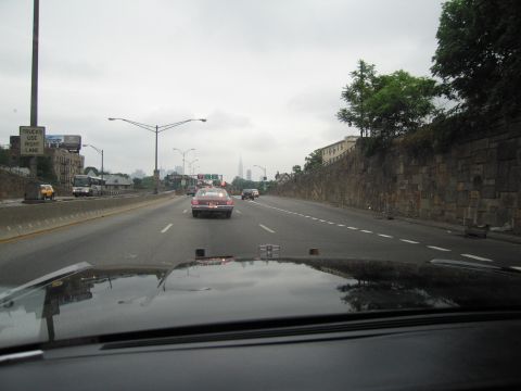 1991 Ford LTD Crown Victoria - maroon car drives ahead on a multi-lane highway with a stone retaining wall and a hazy city skyline in the distance.