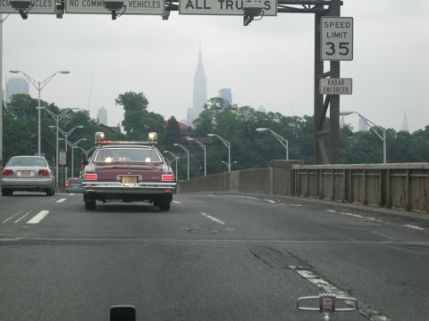 1991 Ford LTD Crown Victoria - maroon station wagon with roof beacons climbs a bridge approach below overhead signs, the Manhattan skyline rising behind.