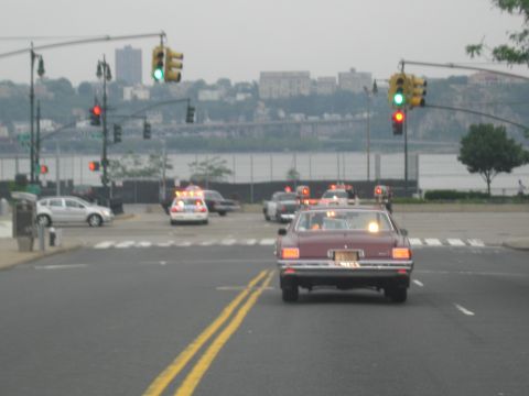 1991 Ford LTD Crown Victoria - maroon sedan with roof beacons crosses an intersection with traffic signals, a river and hills in the background.