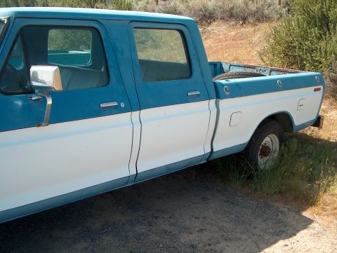 1978 Ford Bronco - Side view of a blue and white two-tone 1970s Ford crew cab long-bed pickup parked on gravel beside dry brush, with a faded paint and a rust…