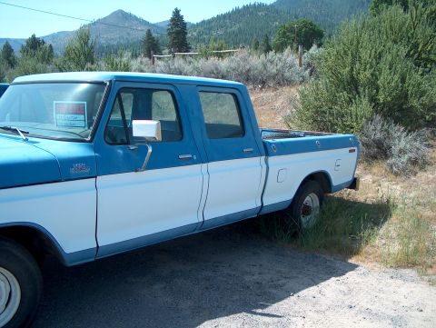 1978 Ford Bronco - Blue and white two-tone 1970s Ford F-250 Custom crew cab long-bed pickup parked on a gravel shoulder with pine-covered hills behind it. A h…