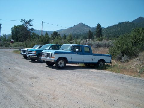 1978 Ford Bronco - Three older Ford trucks lined up nose-out on a dirt shoulder in dry mountain country, seen from the 3/4 front. Closest is a blue-over-white…