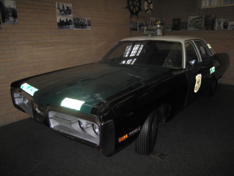 1991 Ford LTD Crown Victoria - vintage dark-green and white police sedan sits on display indoors before a brick wall hung with historical photographs.
