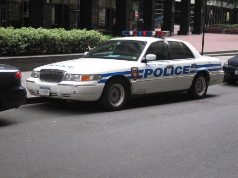 1991 Ford LTD Crown Victoria - white Mercury Grand Marquis police cruiser with blue striping and roof light bar parks along a downtown city street.