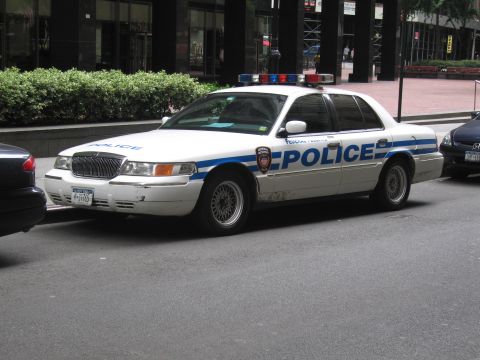 1991 Ford LTD Crown Victoria - white police sedan marked POLICE with a blue stripe and roof light bar sits parked curbside on a city street.