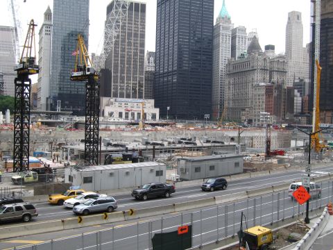 1991 Ford LTD Crown Victoria - Tower cranes rise over an excavated construction site beside a busy multi-lane road backed by tall city buildings.
