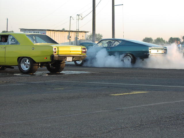 Yellow and teal muscle cars on a drag strip, one doing a burnout