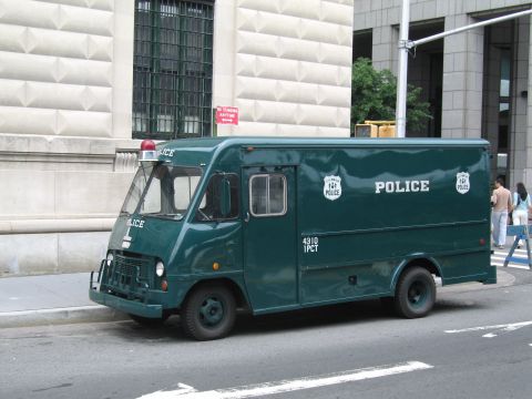 1991 Ford LTD Crown Victoria - dark-green police step van lettered POLICE with a red roof beacon parks along a city street beside a stone building.