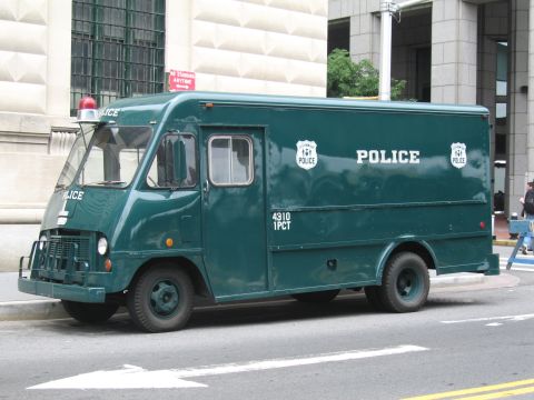 1991 Ford LTD Crown Victoria - Side view of a dark-green police step van lettered POLICE and 4310 1PCT, parked curbside near a stone building.