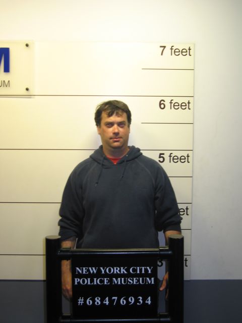 1991 Ford LTD Crown Victoria - man in a gray hooded sweatshirt stands facing forward against a mugshot height chart holding a New York City Police Museum booking placard.