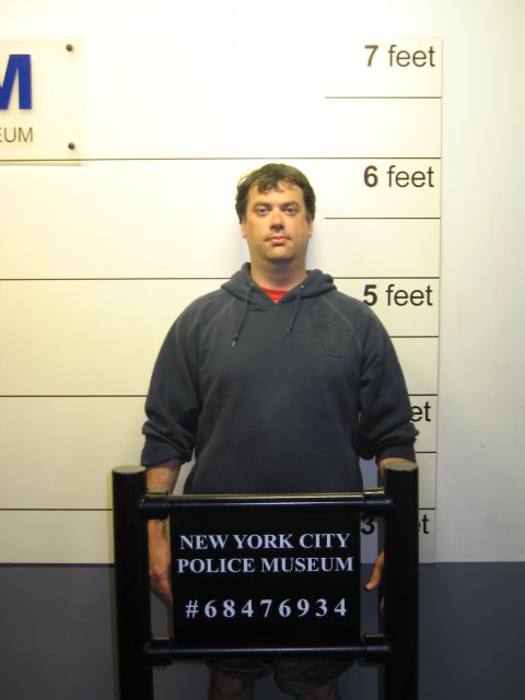1991 Ford LTD Crown Victoria - man in a gray hoodie stands behind a black New York City Police Museum booking placard reading number 68476934 in a mock mugshot setup.