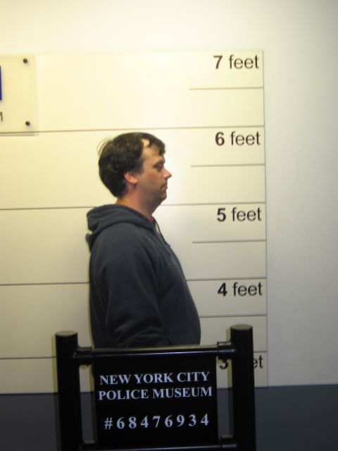1991 Ford LTD Crown Victoria - Side-profile mock mugshot of a man in a gray hoodie against a height chart with a New York City Police Museum placard in front.