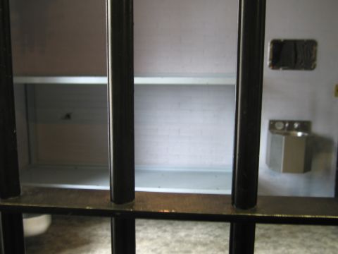 1991 Ford LTD Crown Victoria - Interior of a mock jail cell showing a stainless steel shelf bunk and bare brick wall viewed through vertical black bars.