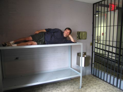 1991 Ford LTD Crown Victoria - man in a dark shirt and shorts lies propped on his elbow atop a steel shelf bunk in a mock jail cell beside a barred gate.