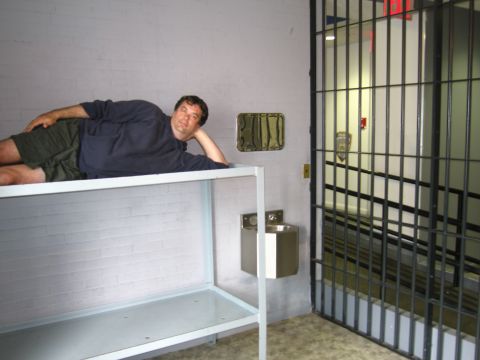 1991 Ford LTD Crown Victoria - man rests on his side on a steel shelf bunk inside a brick-walled mock jail cell with a stainless drinking fountain and barred gate nearby.