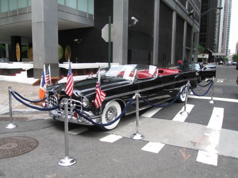 1991 Ford LTD Crown Victoria - long black vintage convertible parade car with red interior, mounted American and city flags, roped off on a city sidewalk.