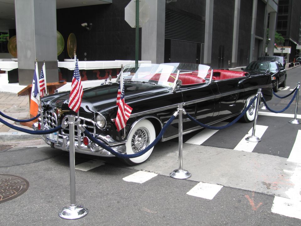 Black vintage convertible parade car with flags roped off by skyscraper