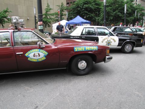 1991 Ford LTD Crown Victoria - maroon Pontiac LeMans in Missouri State Highway Patrol livery sits beside other vintage patrol cars at an outdoor event.