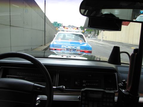 1991 Ford LTD Crown Victoria - Dashboard-level view through a windshield following a blue-and-white POLICE cruiser on a roadway, with the host car's dash and steering whe…