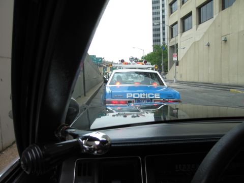1991 Ford LTD Crown Victoria - blue-and-white POLICE cruiser drives ahead on a city road, viewed through the windshield of a following vehicle past its A-pillar.