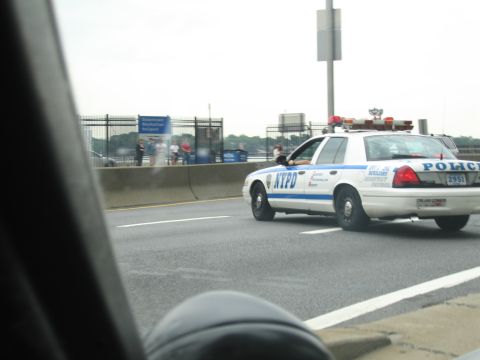 1991 Ford LTD Crown Victoria - white NYPD police car with light bar drives on a highway, photographed from a passing vehicle with a guardrail and fence behind.