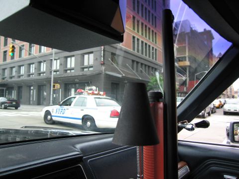 1991 Ford LTD Crown Victoria - white NYPD police car waits at a city intersection, viewed through the side window of a car with a dark dashboard in the foreground.