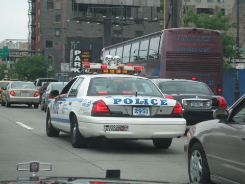 1991 Ford LTD Crown Victoria - white NYPD POLICE car with roof light bar sits in city traffic alongside a maroon tour bus and a black sedan.