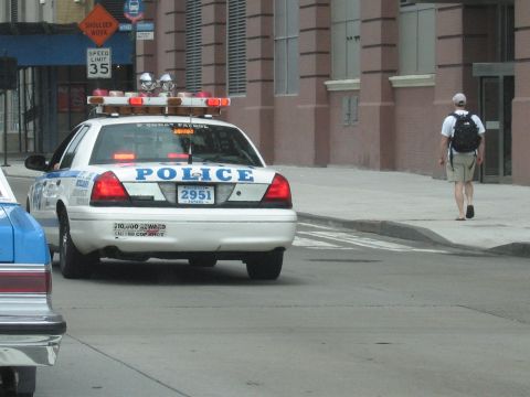 1991 Ford LTD Crown Victoria - white POLICE car with roof light bar idles at a curb on a city street while a man with a backpack walks along the sidewalk.