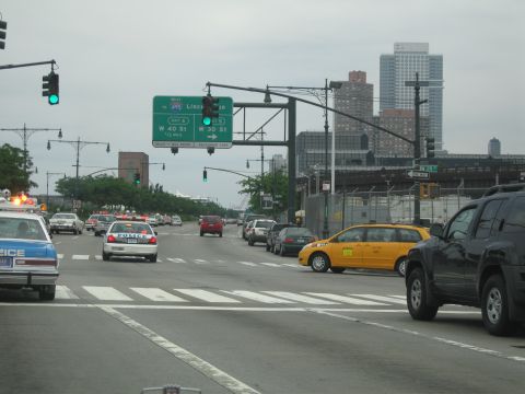 1991 Ford LTD Crown Victoria - broad city avenue with several POLICE cruisers, a yellow taxi and a dark SUV beneath green highway signs for the Lincoln Tunnel approach.