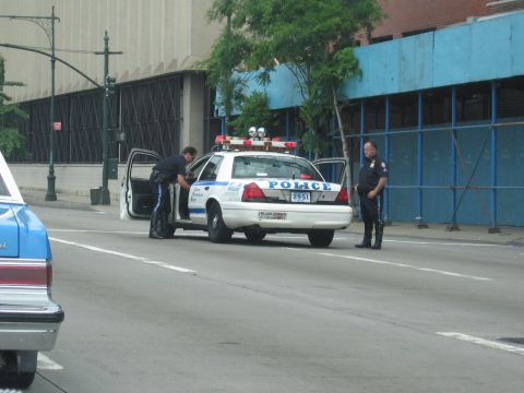 1991 Ford LTD Crown Victoria - Two uniformed officers stand by a white POLICE car with its driver door open on a quiet urban street lined with a blue construction barrier.