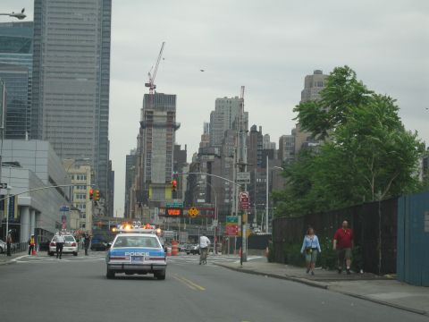 1991 Ford LTD Crown Victoria - white POLICE car drives down a wide city avenue toward a cluster of high-rise buildings, with pedestrians on the sidewalk.