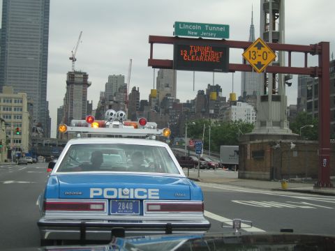 1991 Ford LTD Crown Victoria - blue-and-white Dodge POLICE car with roof lights heads toward the Lincoln Tunnel beneath green and yellow clearance signs and a city skylin…