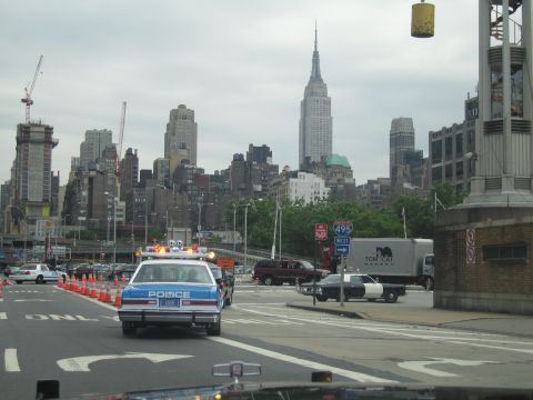 1991 Ford LTD Crown Victoria - blue-and-white POLICE car drives a multilane road past orange cones with a tall city skyline including the Empire State Building behind.
