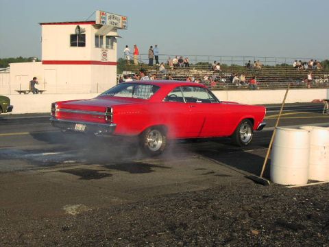 1981 Ford Bronco - A red coupe performs a burnout at a drag strip, with spectators in the background.