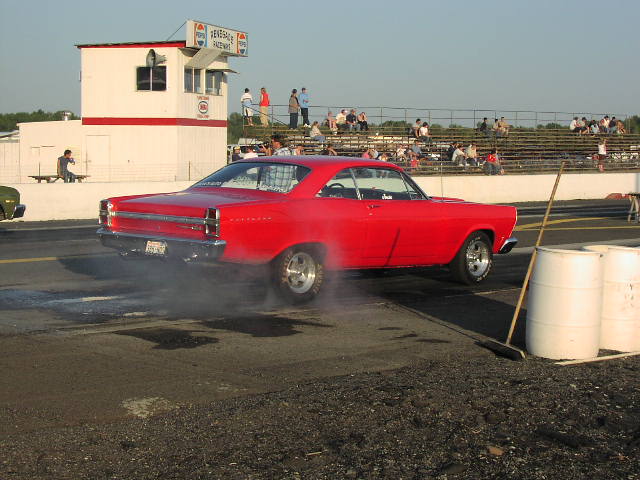 Red coupe with racing slicks, smoking tires, and a roll cage, viewed from the side at a drag strip
